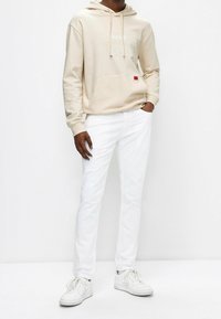 Beige Kapuzenpullover mit einer Fronttasche und weißen Text, kombiniert mit schmal geschnittenen weißen Hosen und weißen Sneakern. Lässiges und modernes Design.