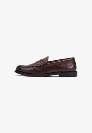 Mocasín marrón de cuero para hombre con suela negra, costura moc toe y pequeño emblema metálico en el lateral superior.