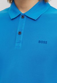 Blå pikétröja i texturerad tyg med krage, två svarta knappar och en diskret präglad logotyp på bröstet.