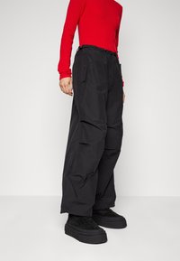 Donna che indossa pantaloni cargo neri larghi, scarpe platform nere e una maglietta rossa aderente a maniche lunghe, su uno sfondo chiaro e semplice.