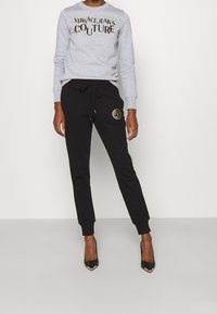 Person wearing grey Versace Jeans Couture sweatshirt and black joggers with gold logo, paired with black pointed-toe heels.