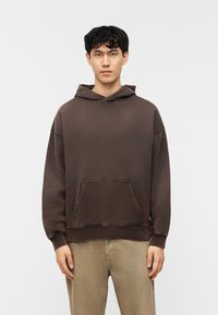 Νέος άνδρας με κοντά σγουρά μαύρα μαλλιά φοράει καφέ φαρδιά φούτερ και μπεζ παντελόνι, στέκεται μπροστά σε απλό λευκό φόντο.