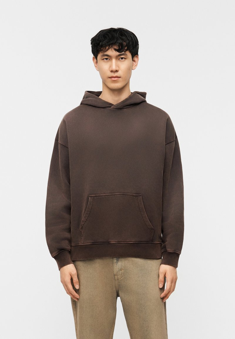 Νέος άνδρας με κοντά σγουρά μαύρα μαλλιά φοράει καφέ φαρδιά φούτερ και μπεζ παντελόνι, στέκεται μπροστά σε απλό λευκό φόντο.