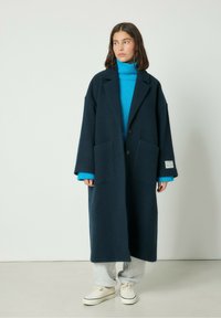 Námornícky vlnený kabát s uvoľneným strihom, dvomi prednými vreckami a zapínaním na gombík, vrstvený nad živou modrou rolákou a šedými teplákmi.