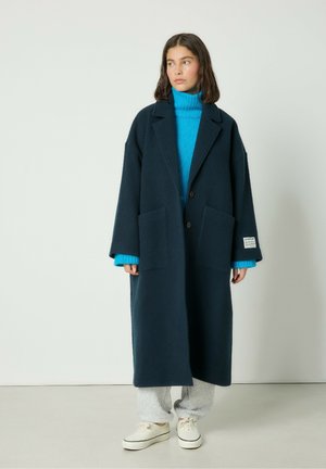 Marineblå uldblandingsfrakke med afslappet pasform, to forlommer og knaplukning, lagt over en livlig blå turtleneck og grå sweatpants.