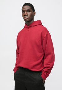 Felpa con cappuccio rossa dal taglio comodo, polsini a costine e piccolo logo sul petto. Abbinata a pantaloni neri, sfondo semplice.