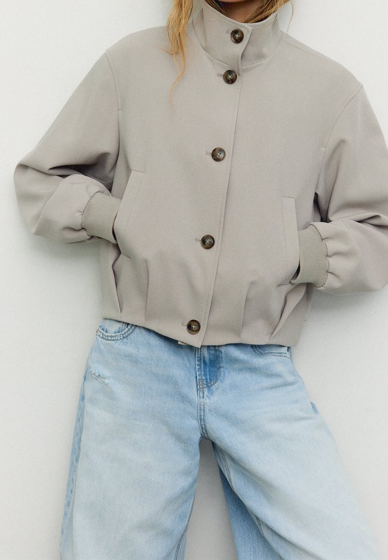 Personne portant une veste beige boutonnée avec un col montant et des poignets élastiques, associée à un jean en denim bleu clair taille haute.
