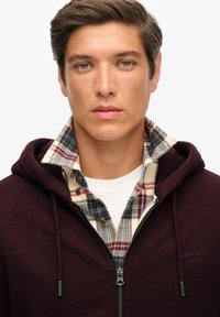 Haut à capuchon zippé bordeaux avec une finition texturée, superposé à une chemise à carreaux comportant des tons de beige, bleu et rouge.