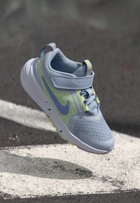 Light blue athletic shoe with mesh upper, contrasting yellow and green accents, and a Velcro strap. Features a textured sole for grip.