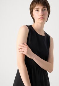 Crna haljina bez rukava s okruglim izrezom, uskim korzetom i lepršavom suknjom. Glatka tekstura s minimalnim dizajnerskim detaljima.