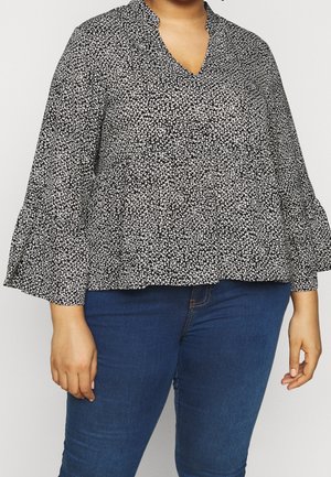 Kobieta ubrana w czarno-białą kwiecistą bluzkę z długim rękawem i falbankami na rękawach oraz niebieskie dżinsy, widoczna od szyi do bioder.