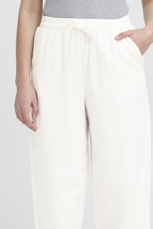 Gros plan d'une personne portant un pantalon blanc à cordon avec taille élastique et un haut gris sans manches, main reposant dans la poche.