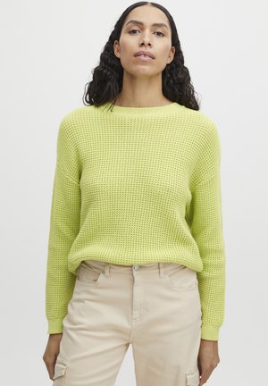 Brede trui in fel limoen groen met een gestructureerd patroon, ronde halslijn en lange mouwen. Gecombineerd met beige broeken.