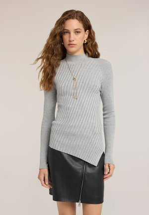 Kvinde med langt krøllet hår iført en grå ribstrikket sweater og sort lædernederdel med lynlås foran, stående mod en ensfarvet baggrund.