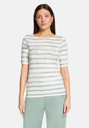 Kvinde iført kortærmet top med hvide og lysegrønne striber og abstrakt prikket design, kombineret med lysegrønne bukser, stående foran en ensfarvet baggrund.