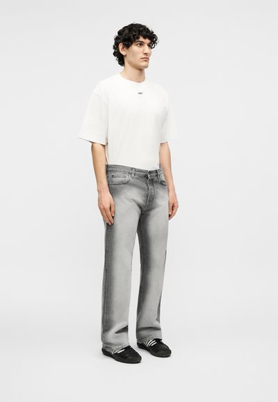 Νεαρός άνδρας όρθιος, φοράει λευκό φαρδύ T-shirt, ξεβαμμένα γκρι τζιν με ίσια γάμπα και μαύρα αθλητικά παπούτσια με άσπρες ρίγες.