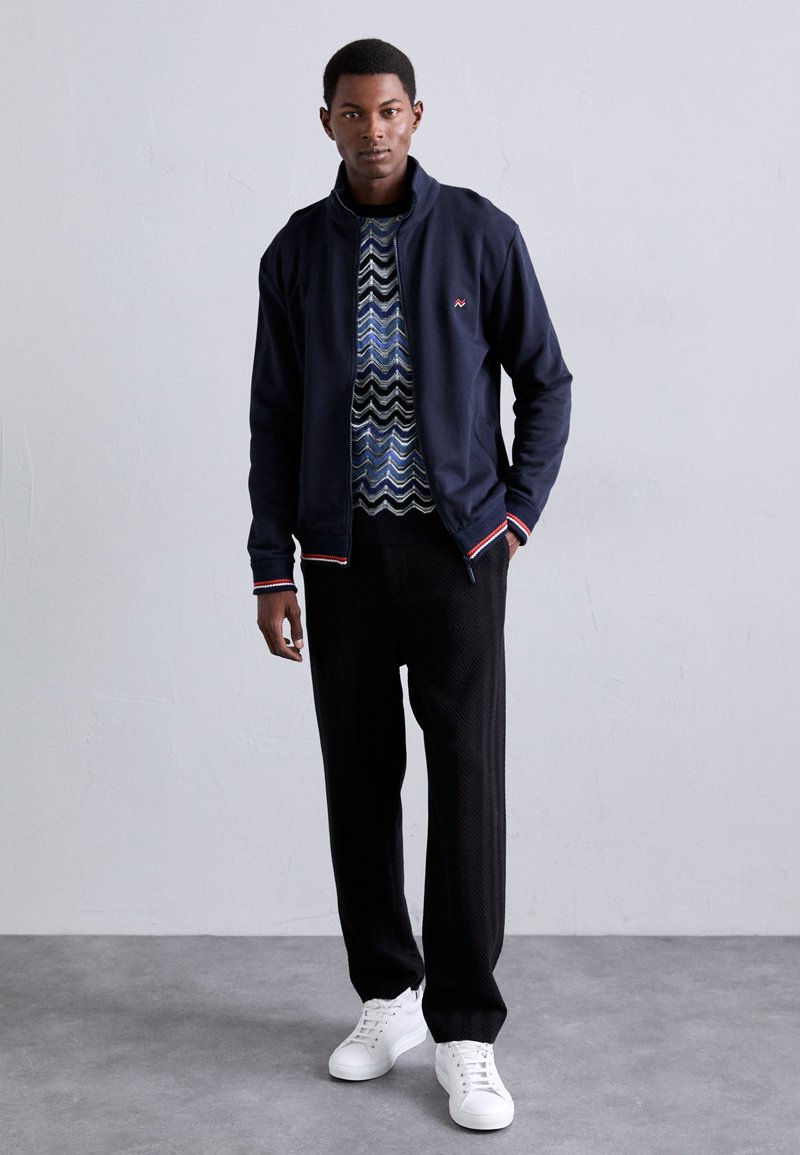 Navy Zip-Jacke mit roten und weißen Streifenakzenten, gemustertem Pullover darunter, strukturierten schwarzen Fischgrät-Hosen und weißen Sneakern.