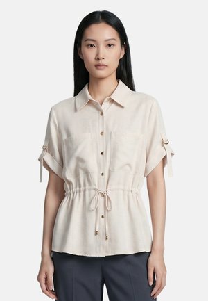 Donna che indossa una camicetta beige chiaro a maniche corte con bottoni, vita regolabile con coulisse e maniche arrotolate, in piedi davanti a uno sfondo semplice.