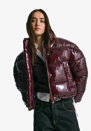 Burgundy glossy puffer jacket with a boxy shape, zip closure, and visible stitching details. Worn over a black sweater and white shirt.
