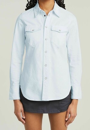 Femme portant une chemise bleu clair à boutons avec deux poches poitrine et une mini-jupe sombre, debout devant un fond uni.