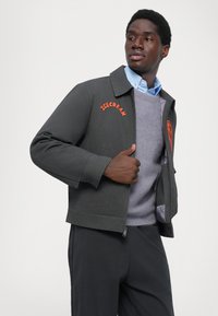 Grijze jas met oranje "ICECREAM" borduursel, ritssluiting en twee voorzakken. Gedragen over een lichtbl blouse en een grijze trui.