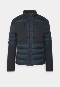 Marinblå puffjacka med en slät yta, dragkedjefickor på bröstet och hög krage. Har kviltade sektioner och åtsittande ärmar.