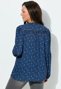 Marineblauwe blouse met lichtblauw hartjespatroon, lange mouwen, geplooide achterkant en subtiele plooiing langs de inzet. Zachte stoftextuur.