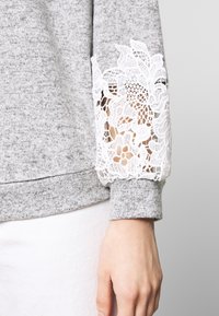 Grå sweatshirt med vita spetsdetaljer på ärmarna. Har ett mjukt, texturerat tyg och ribbade ärmslut.