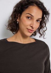 Mujer con cabello rizado y corto de color oscuro, usando pendientes de aro de plata y una blusa marrón oscuro, sonriendo suavemente frente a un fondo liso.