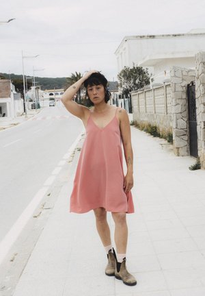 Różowa sukienka bez rękawów z dekoltem w serek, wykonana z gładkiej tkaniny. W połączeniu z brązowymi botkami za kostkę i białymi skarpetkami, stojąca na chodniku.