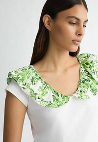 T-shirt blanc avec un col à volants à motif floral vert, présentant une encolure ronde et des manches courtes. Texture de tissu lisse.