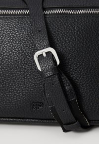 Sac en cuir noir avec une finition texturée, doté d'une fermeture éclair en métal argenté et d'une boucle de sangle. Comprend un détail de logo sur le devant.