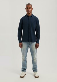 Marineblauwe longsleeve shirt met een gestructureerd patroon, met knopen aan de voorkant, een enkele borstzak, gecombineerd met vervaagde lichtblauwe jeans en witte sneakers.