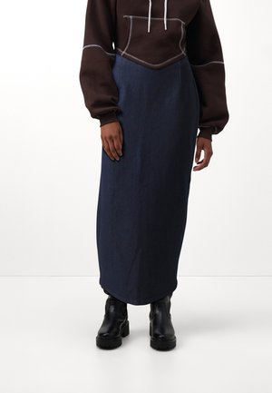 Personne portant un sweat à capuche marron foncé, une longue jupe bleu marine et des bottines noires épaisses, debout devant un fond blanc uni.