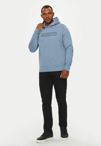 Felpa blu realizzata in tessuto morbido con tasca a marsupio e cappuccio con cordino. Presenta un logo grigio "Calvin Klein" sul petto, abbinato a pantaloni neri.