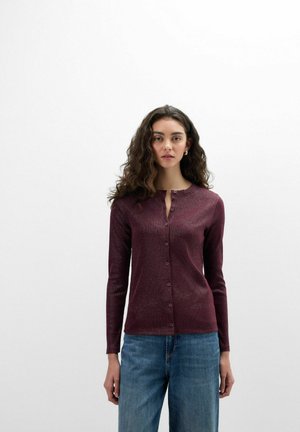 Frau mit langen lockigen Haaren, die eine burgunderfarbene, geknöpfte Strickjacke und blaue Jeans trägt, steht vor einem einfarbigen hellen Hintergrund.