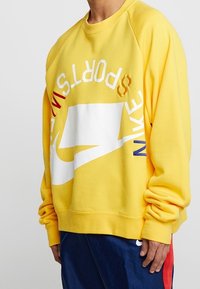 Felpa gialla con vestibilità oversize, caratterizzata da un logo Nike bianco e testo multicolore. Realizzata in materiale morbido con polsini a coste.