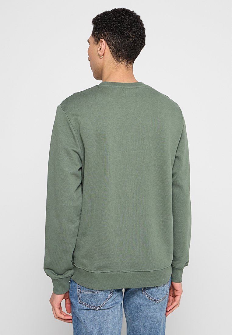 Sudadera verde olivo con cuello redondo, mangas largas y puños acanalados. La tela parece suave con un acabado texturizado, llevada sobre jeans azul claro.