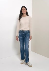 Hellbeige Langarmshirt kombiniert mit lockeren blauen Jeans. Das Model trägt weiße Sneakers mit hellbraunen Akzenten. Einfache, flache Texturen.