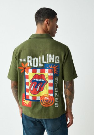 Mann trägt olivgrünes kurzärmeliges Hemd mit farbigem Rolling-Stones-Logo und Text auf der Rückseite, steht vor einem einfarbigen Hintergrund.