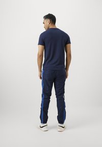 Homem de pé, de costas, a usar uma t-shirt azul marinho, calças de treino azul marinho com riscas azuis e ténis brancos com logótipos pretos de swoosh.