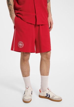 Mężczyzna ubrany w czerwone sportowe szorty i pasującą koszulkę, białe skarpetki oraz białe trampki z niebieskimi i czerwonymi akcentami, stoi z rękami w kieszeniach.