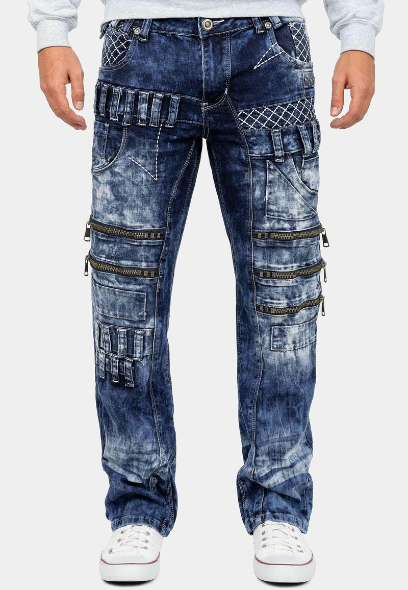 Pantaloni cargo in denim blu scuro con un finish sbiadito, caratterizzati da molteplici tasche con zip e motivi di cucitura decorativa. Dettagli bianchi delle sneaker.