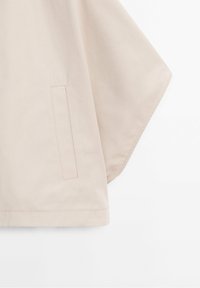 Tissu beige avec une ouverture de poche rectangulaire cousue et une large manche, vu sur un fond blanc.