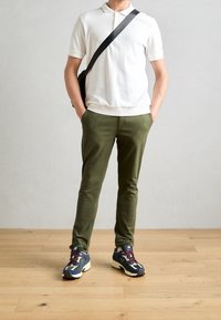 Wit poloshirt, olijfgroene slim-fit broek en een zwarte crossbodytas. Multicolor sneakers met een gestructureerde zool; houten vloer als achtergrond.