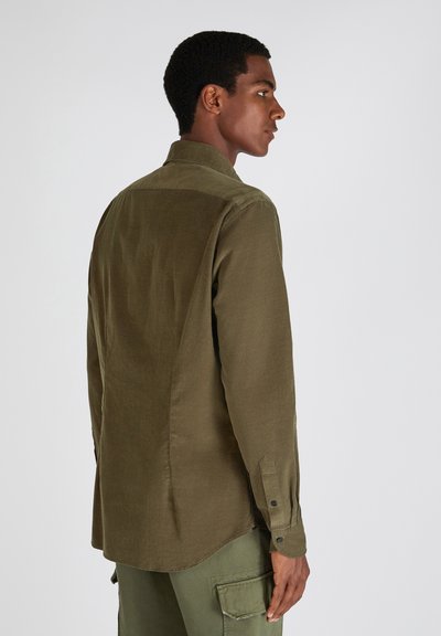 Uomo che indossa una camicia verde oliva con bottoni e pantaloni cargo, in piedi di profilo su uno sfondo chiaro e semplice.
