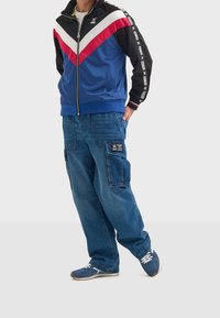 Personne portant une veste de survêtement bleue, rouge, blanche et noire, un jean cargo bleu et des baskets bleues, debout avec une main dans la poche.