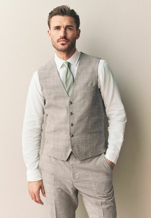 Rövid hajú, szakállas férfi világosszürke kockás mellényben és nadrágban, fehér inggel és világoszöld nyakkendővel, egyik kezével a zsebében áll.