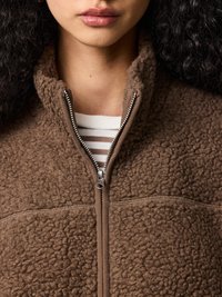 Közeli kép egy személynél, aki barna, texturált cipzáras pulóvert visel fehér és barna csíkos ing felett.