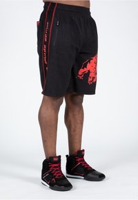 Schwarze, texturierte Shorts mit roten Seitenstreifen und einem roten Grafikdruck. Kombiniert mit schwarzen High-Top-Schuhen mit roten Akzenten und Schnürsenkeln.